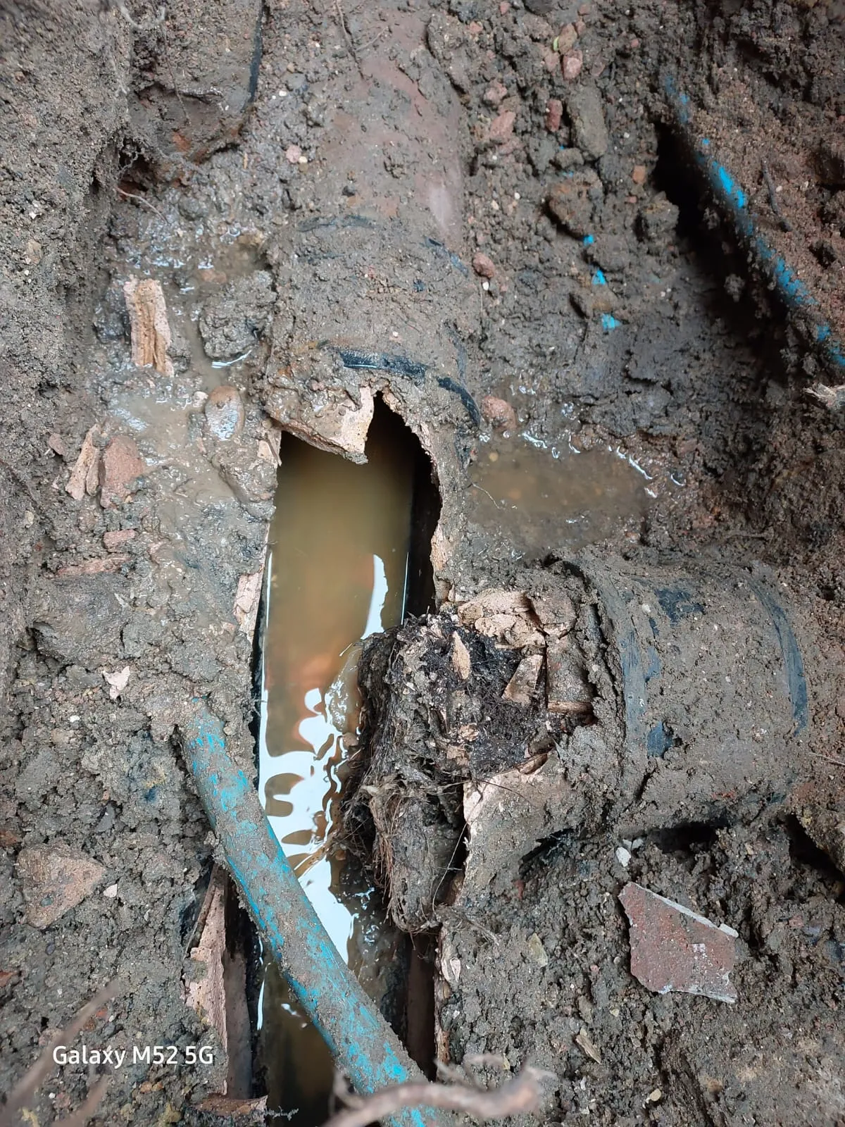 Severely damaged underground drain pipe with muddy water and debris in an excavated trench.