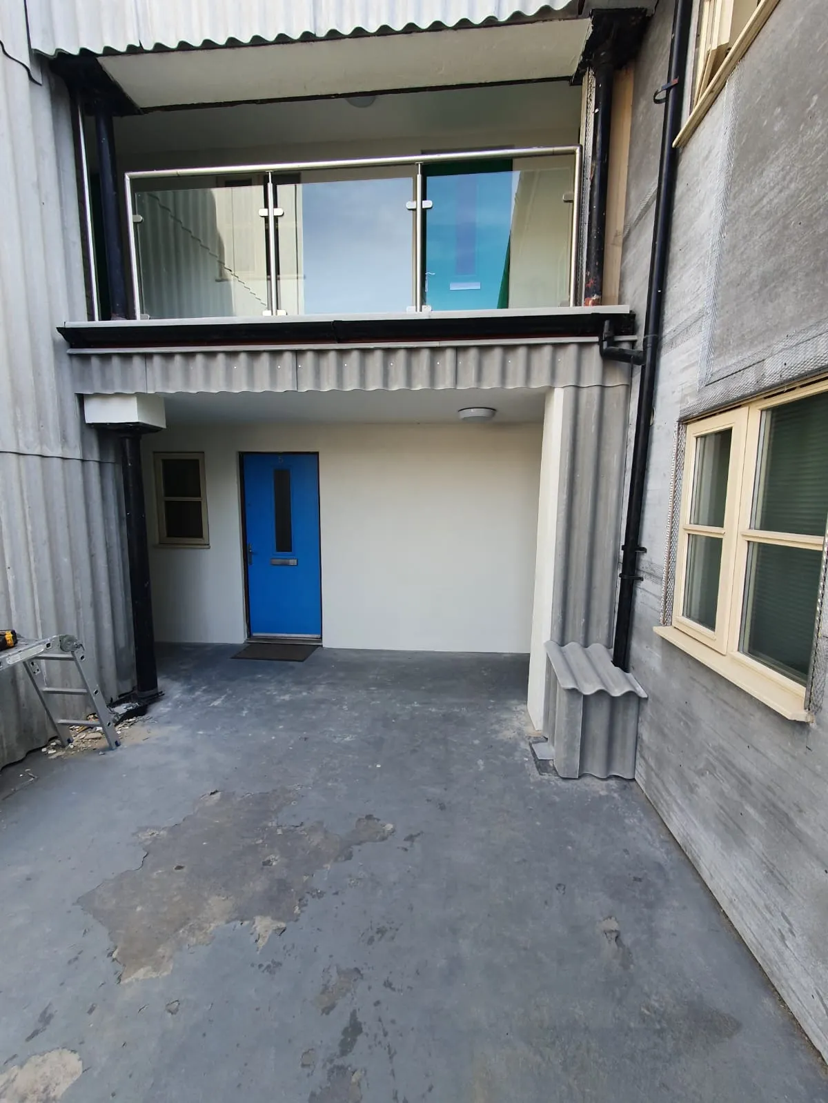 Modern building entrance with a blue door, corrugated overhang, glass balcony, and a visible black downpipe, highlighting the property's roofline and drainage elements.