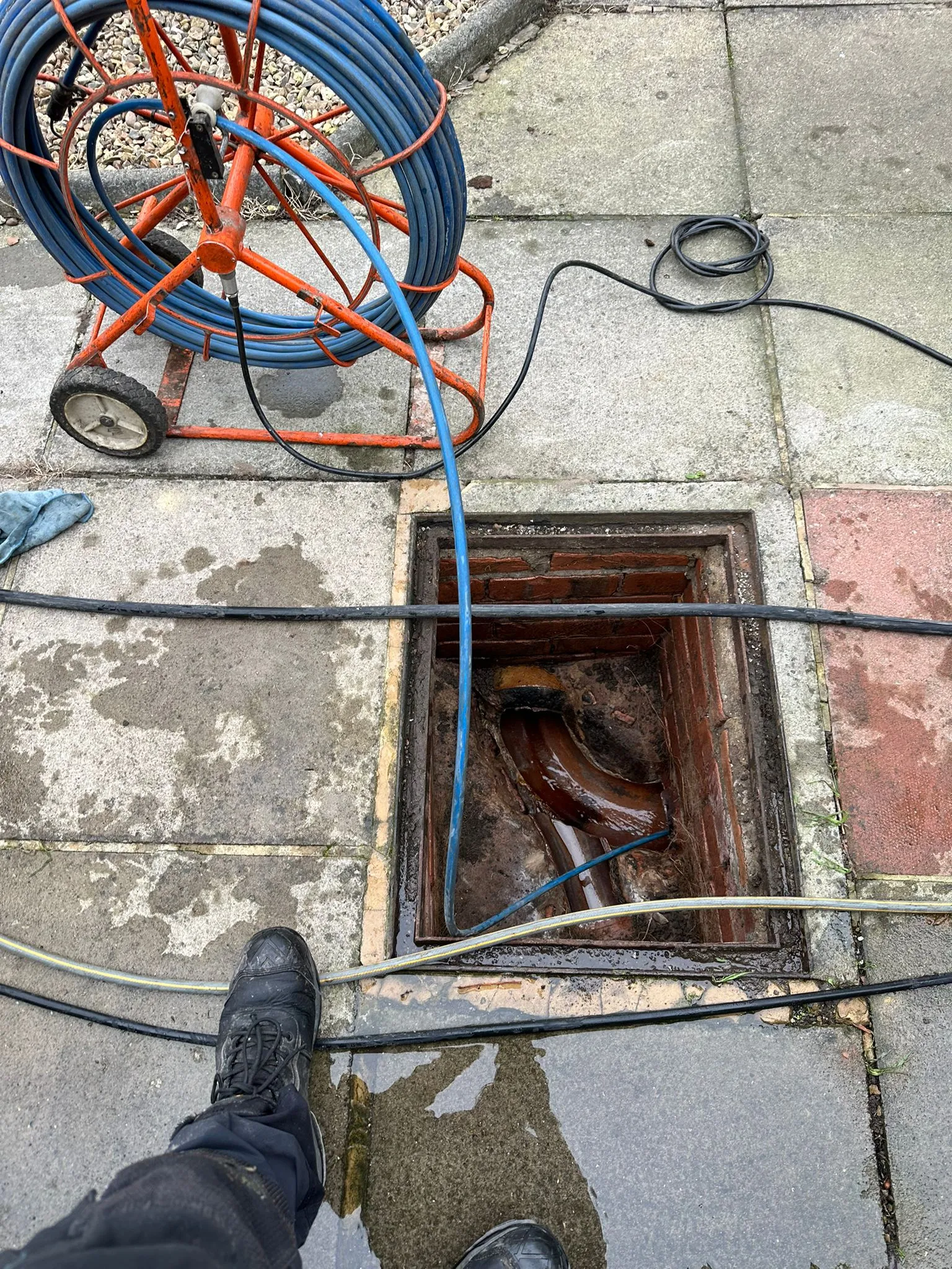 Overhead view of a drainage engineer's boots and an open manhole, with blue high-pressure water jetting equipment on a reel feeding a hose into the drain.