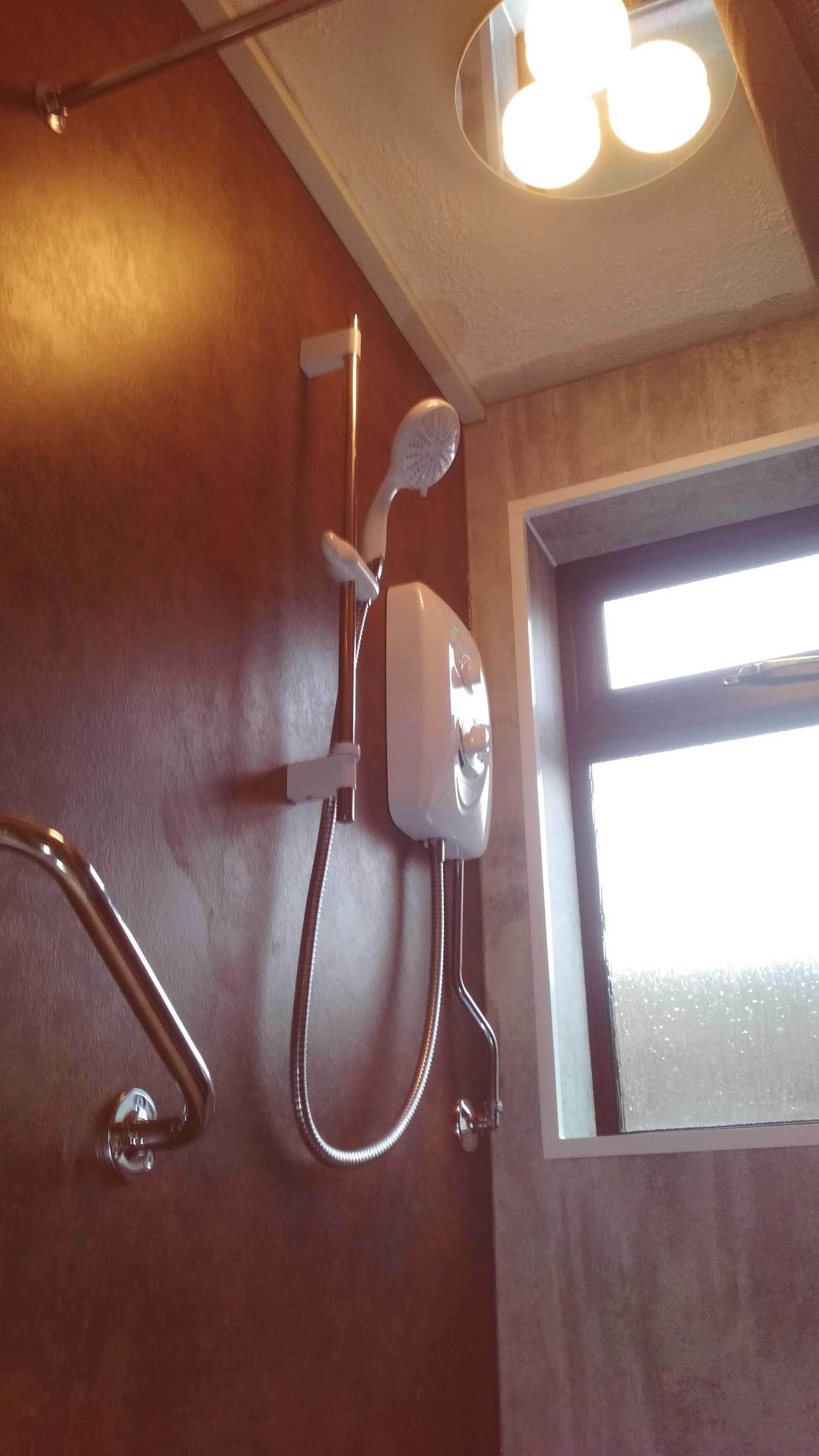 An electric shower unit and shower head installed on a tiled wall in a bathroom, with a grab rail and window visible.