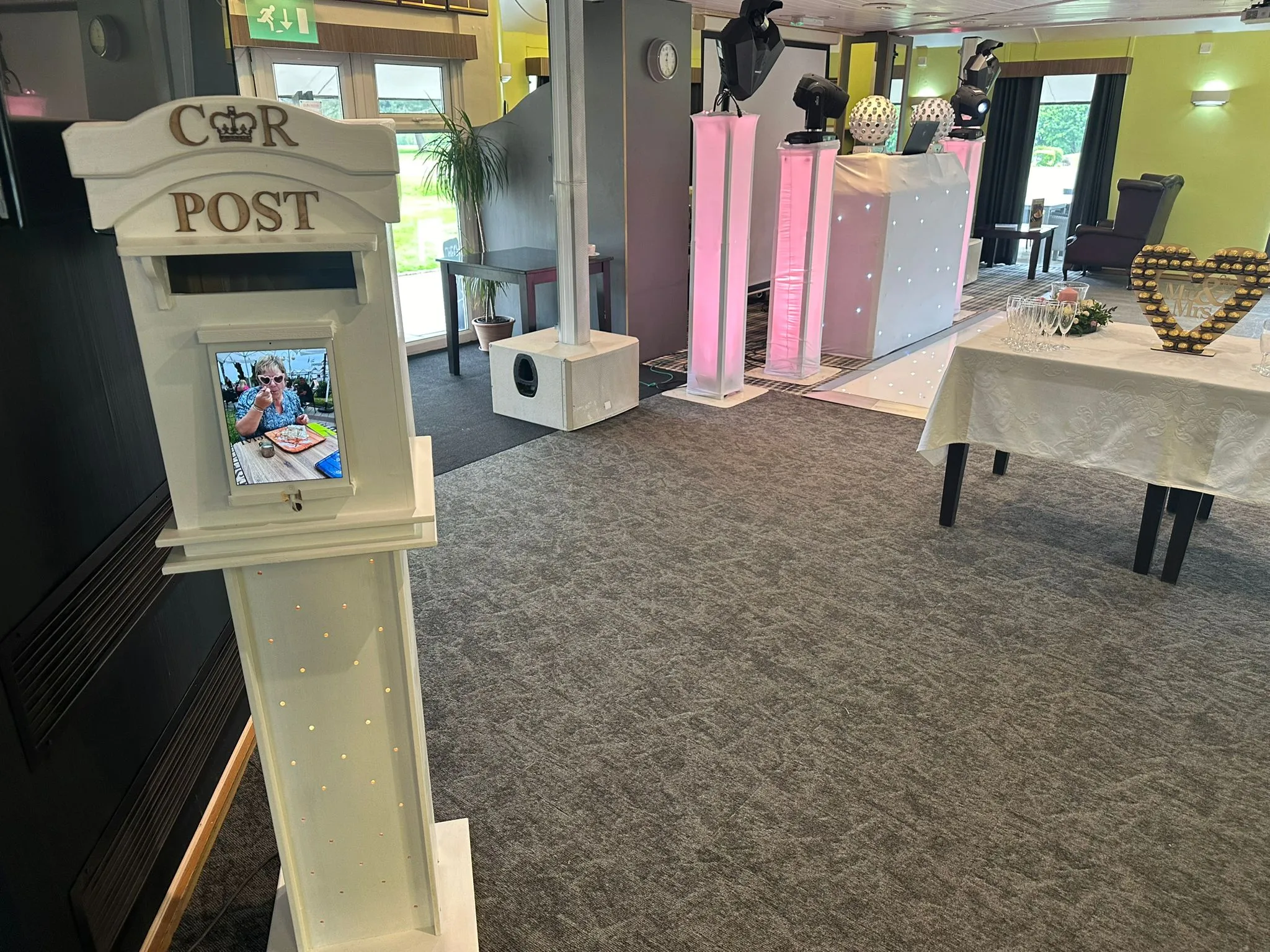 Professional wedding DJ setup with illuminated booth, speakers, ambient pink lighting, a white wedding post box, and a 'Mr & Mrs' heart-shaped chocolate display.