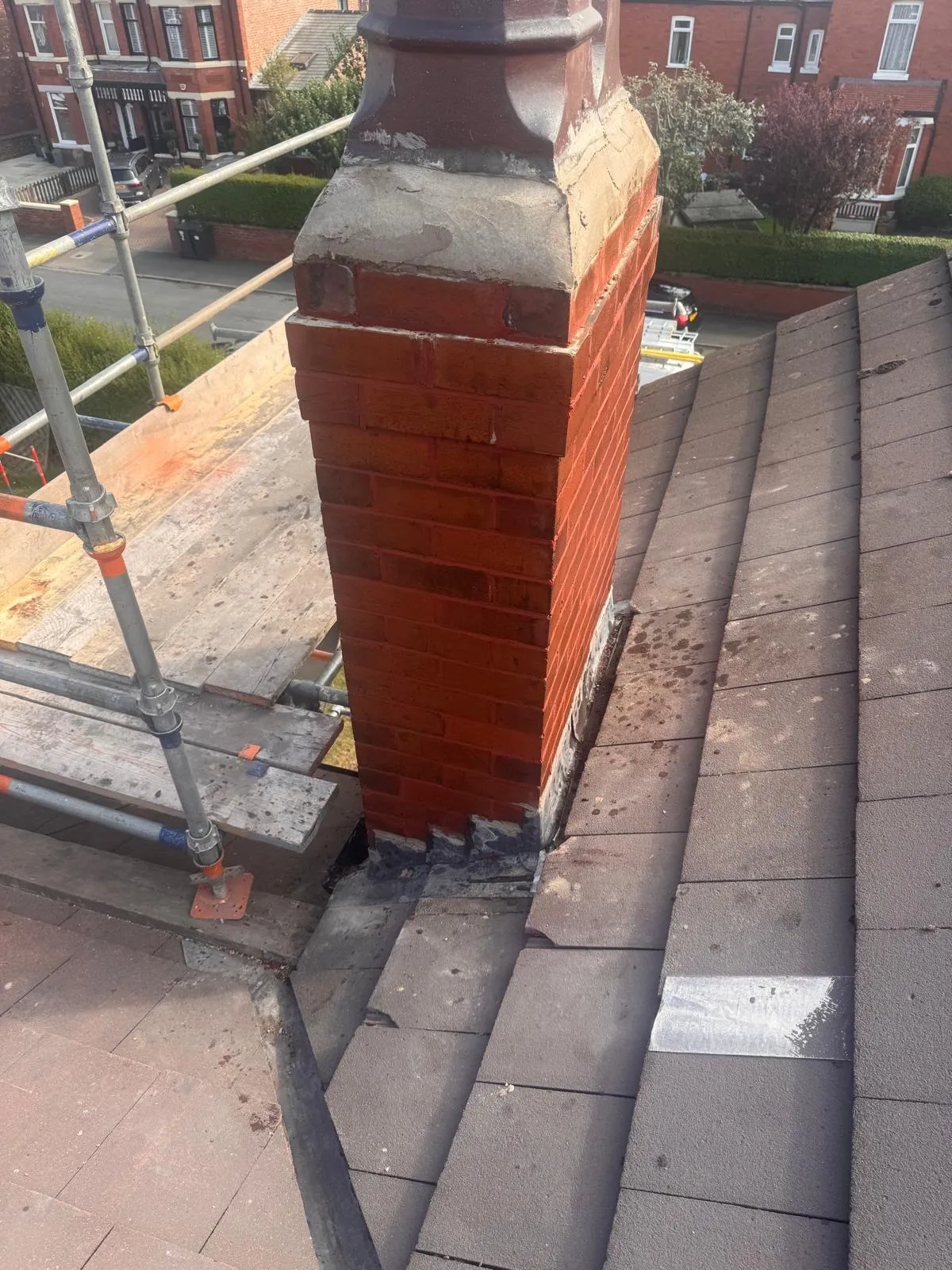 A detailed shot of a red brick chimney on a grey tiled roof, with scaffolding planks and poles visible next to it. The lead flashing around the base of the chimney is prominent.