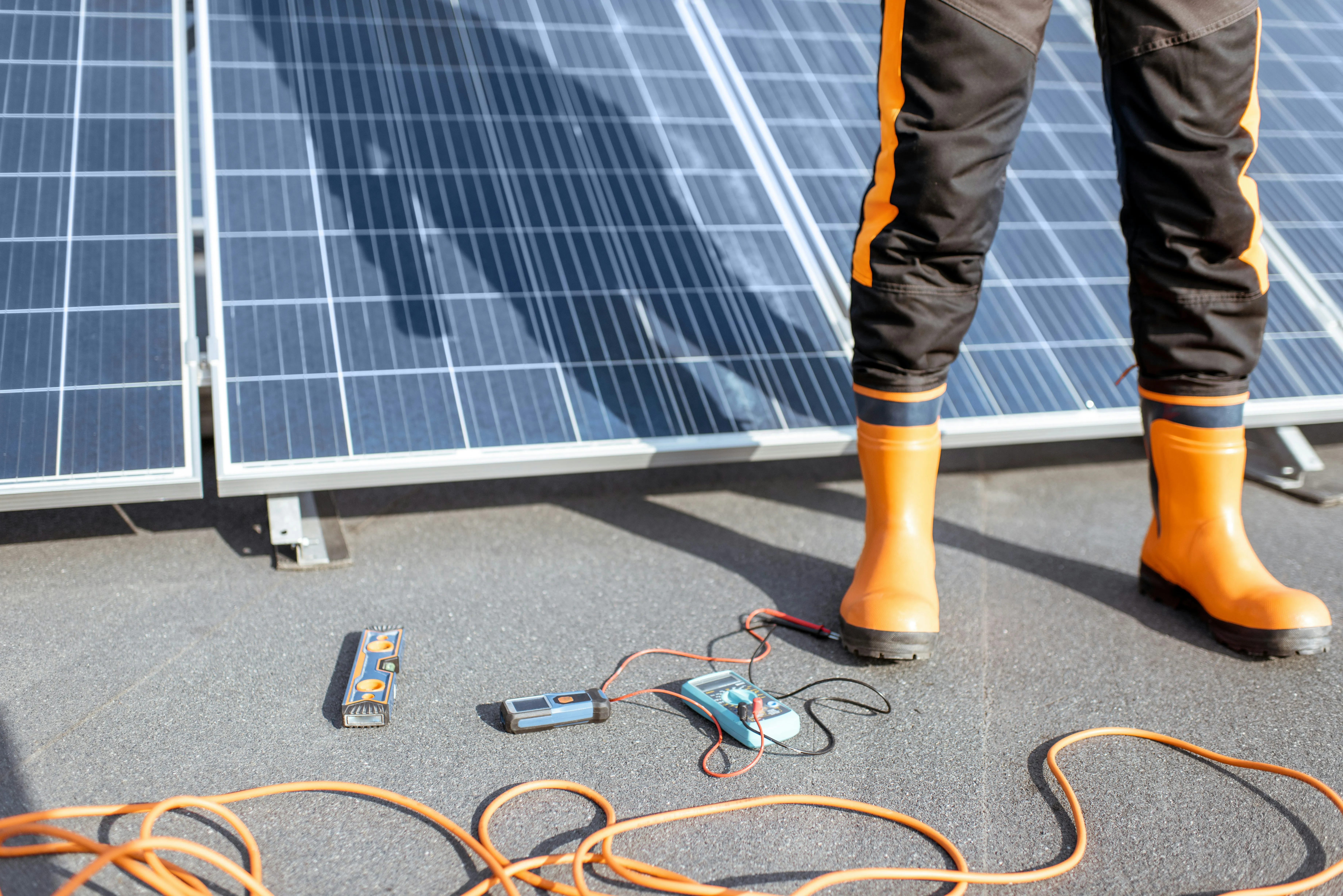 A professional roofer on a flat roof, inspecting solar panels and the roof structure with specialist tools like a multimeter and spirit level, indicating a thorough assessment of the modern roofing system.