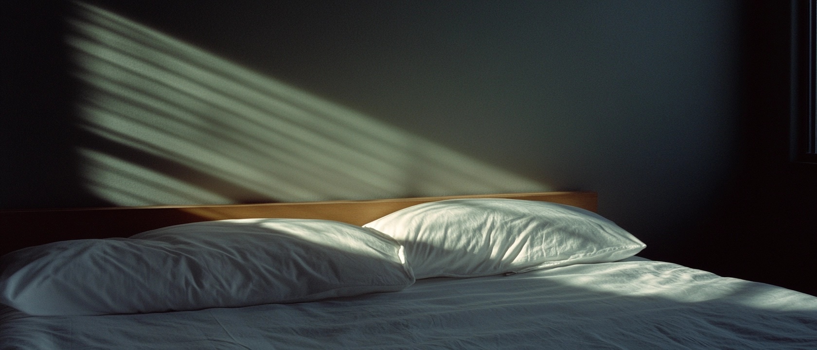 A serene and calming scene of a bed bathed in soft sunlight. The white pillows and sheets create a peaceful atmosphere, while gentle shadows cast by window blinds add depth to the composition. The muted color palette enhances the sense of tranquility and comfort, making it an inviting place for rest and relaxation.