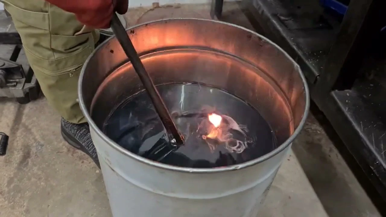 Hot blade plunged into the oil quench bucket, single yellow flame on the surface