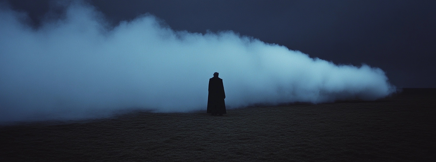 A solitary figure stands in a foggy landscape, shrouded in darkness and illuminated by a mysterious blue haze. The scene evokes feelings of isolation and intrigue, with a backdrop of undefined space disappearing into thick mist. The overall atmosphere is both eerie and contemplative, inviting exploration of the unknown.