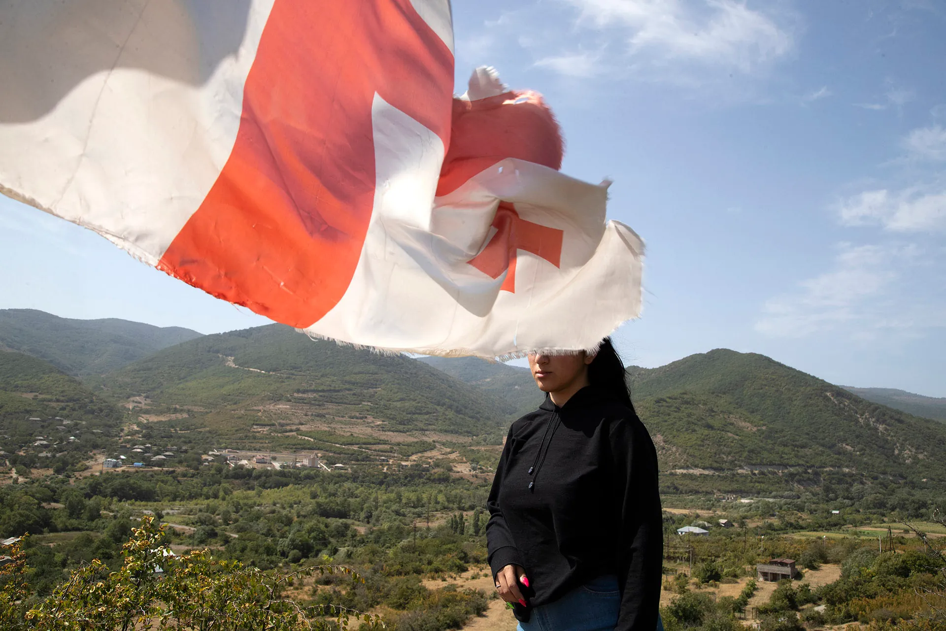 Une femme habillée en noir se tient devant des montagnes. Le drapeau de la Géorgie flotte dans l'air et lui cache les yeux.