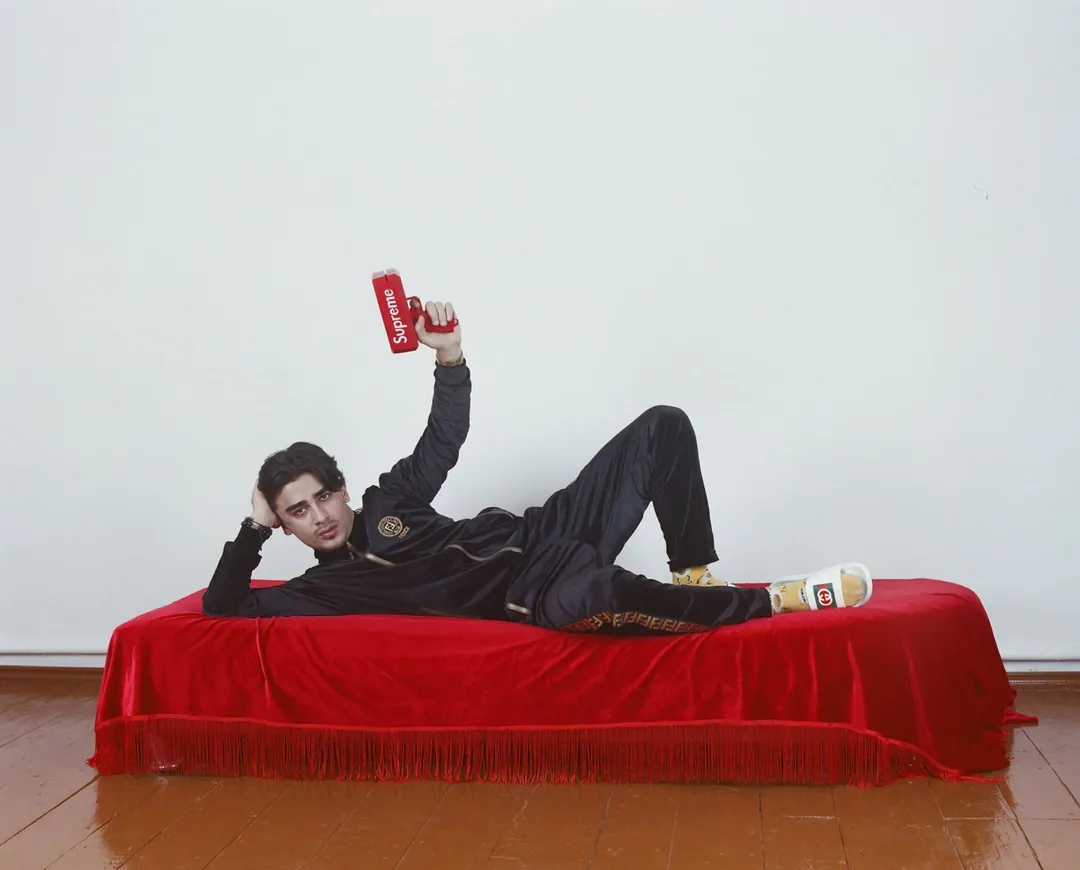 Un homme allongé sur une banquette, avec un pistolet distributeur de billet de la marque Supreme en main, des claquettes Gucci et un ensemble de survêtement Fendi. 
